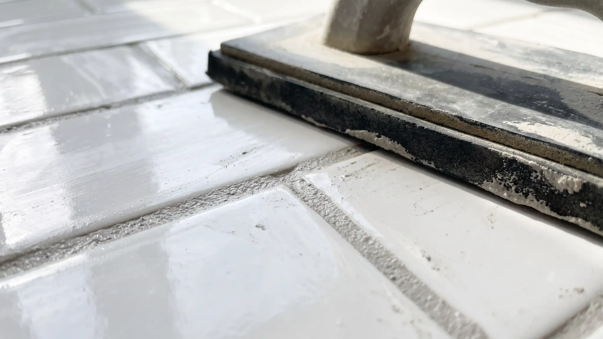 An extreme close view of freshly applied light gray grout lines between white glossy subway tile with a rubber grout float resting on the tile