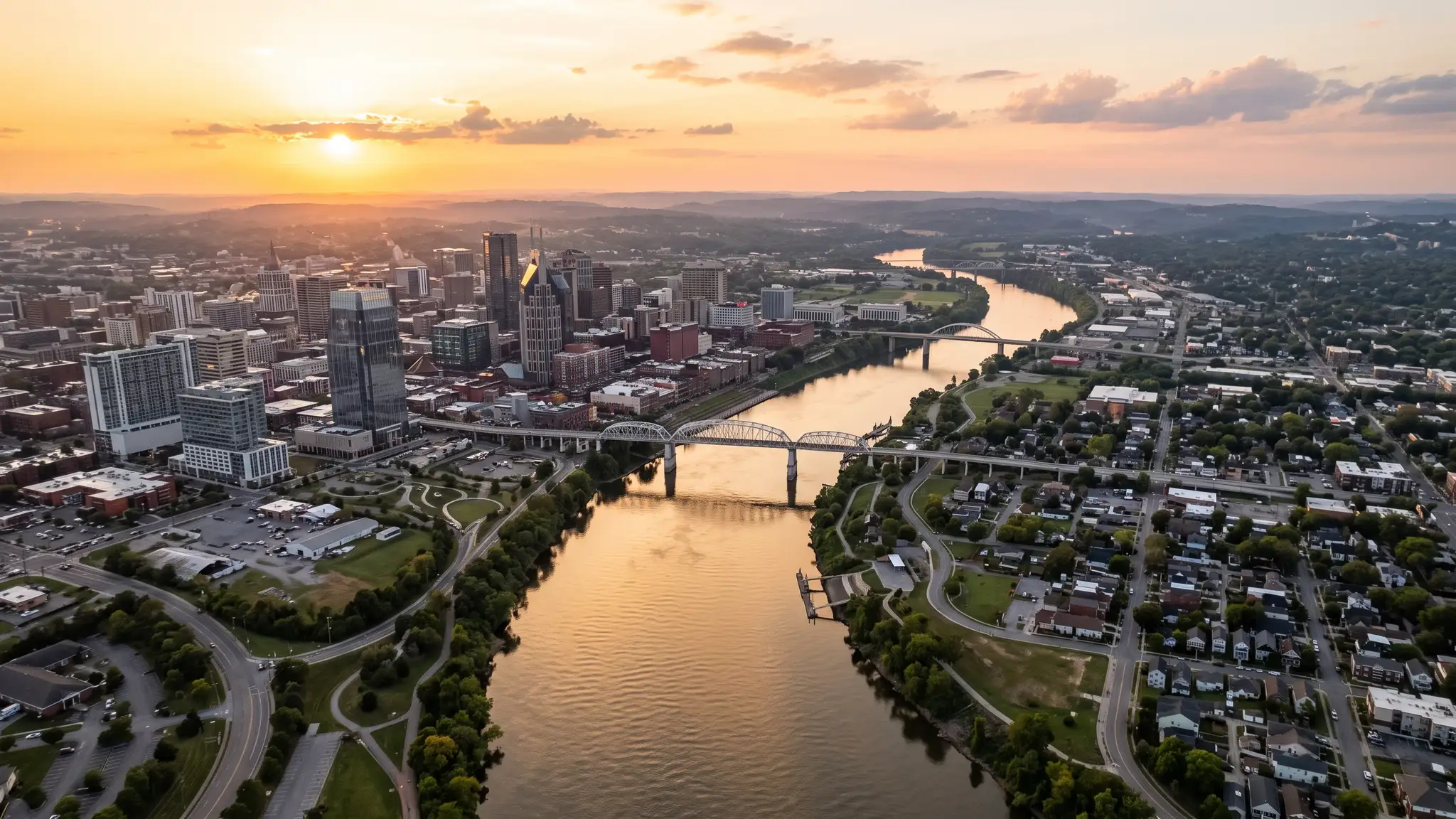 Aerial view of the Cumberland River winding past the Nashville skyline and residential neighborhoods at sunset with golden light on the water